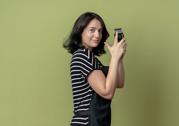 Young beautiful female hairdresser in apron holding hair cutting machine standing sideways  lookign confident over light wall