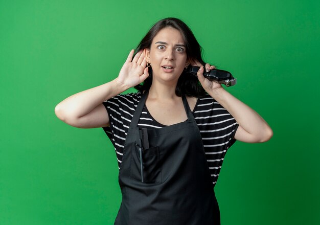 Young beautiful female hairdresser in apron holding hand near ear trying to hear gossip  over green