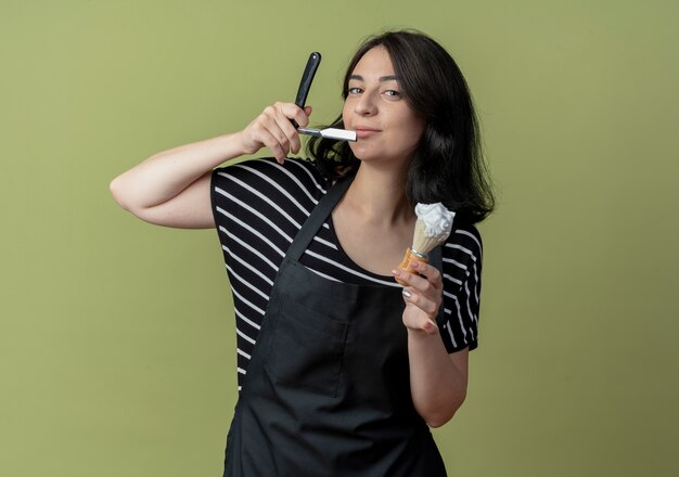 Young beautiful female hairdresser in apron holding razor and shaving brush with foam  smiling confident standing over light wall