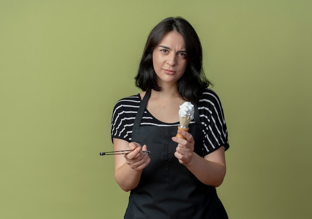 Free photo young beautiful female hairdresser in apron holding razor and shaving brush with foam  with serious face standing over light wall