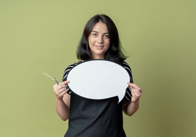 Young beautiful female hairdresser in apron holding scissors blank speech bubble sign  smiling confident standing over light wall