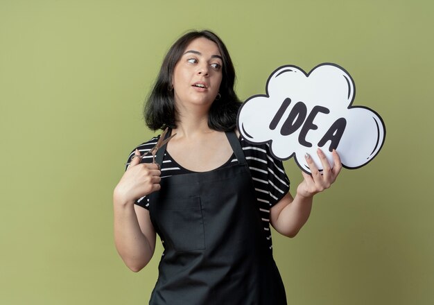 Young beautiful female hairdresser in apron holding scissors and blank speech bubble sign with idea word looking aside confused standing over light wall