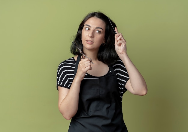 Young beautiful female hairdresser in apron holding scissors combing her hair lookign confident standing over light wall