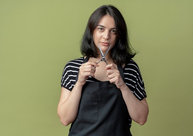 Young beautiful female hairdresser in apron holding scissors smiling confident  over light