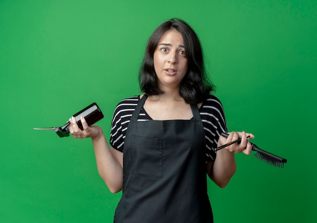 Young beautiful female hairdresser in apron holding scissors spray and brush confused standing over green wall