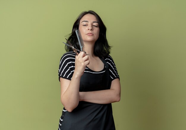 Free photo young beautiful female hairdresser in apron holding scissors with comb looking at them with serious face standing over light wall