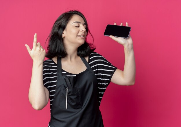 ピンクの壁の上に立っている人差し指を示すスマートフォンを保持しているエプロンの若い美しい女性美容師