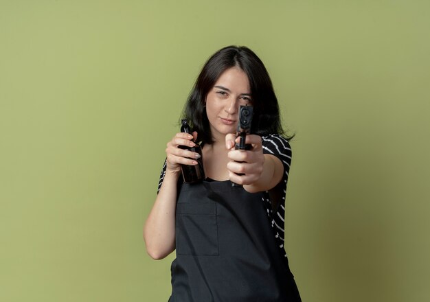 Young beautiful female hairdresser in apron holding spray and aiming gun with serious face standing over light wall