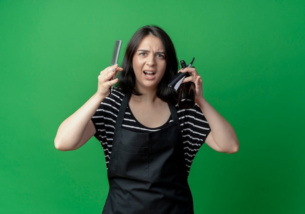 Young beautiful female hairdresser in apron holding spray comb and razor displeased and frustrated  over green