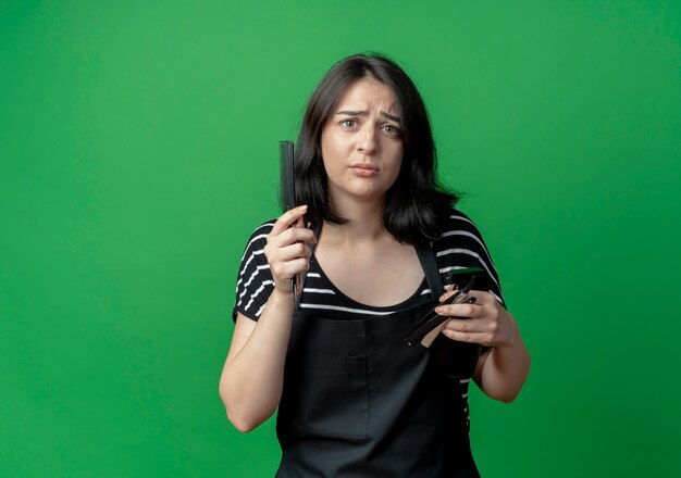 Young beautiful female hairdresser in apron holding spray comb and razor  with arm out as asking question standing over green wall