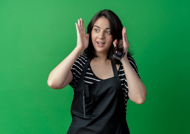 Young beautiful female hairdresser in apron holding trimmer looking aside confused standing over green wall