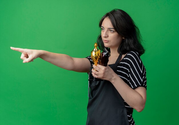 Young beautiful female hairdresser in apron holding trophy and trimmer pointing with finger to the side with skeptic expression  over green