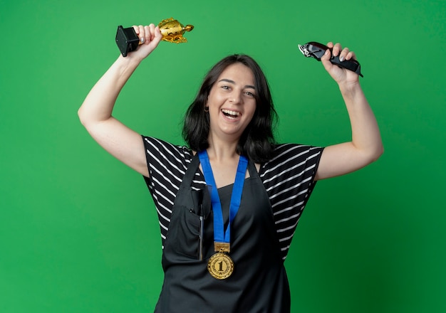 Young beautiful female hairdresser in apron holding trophy and trimmer smiling cheerfully ahppy and excited  over green