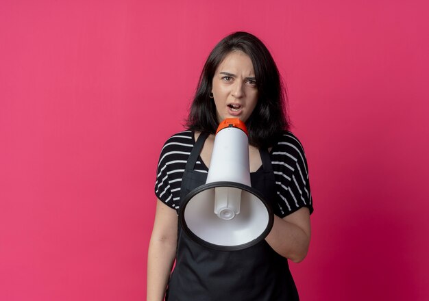 Young beautiful female hairdresser in apron shouting to megaphone standing over pink wall