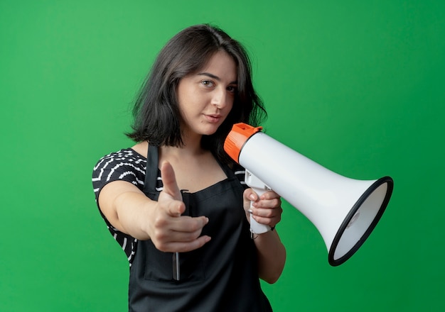 Young beautiful female hairdresser in apron speaking to megaphone pointing with finger at camera looking confident  over green