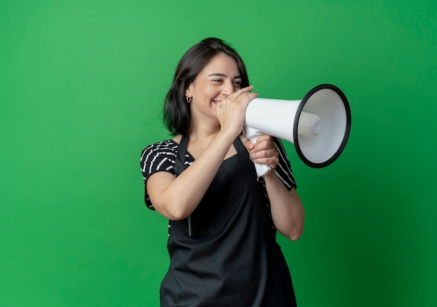 Young beautiful female hairdresser in apron speaking to megaphone with happy face standing over green wall