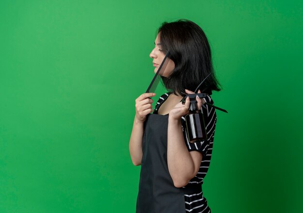 Молодая красивая женщина-парикмахер в стойке перед фартуком держит расческу и бритву над зеленой стеной