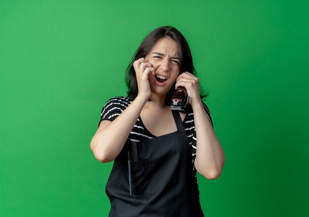 Young beautiful female hairdresser in apron talking on mobile phone with annoyed expression  over green