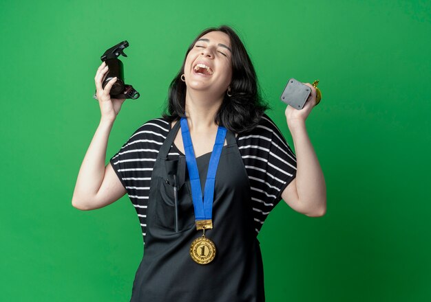 Young beautiful female hairdresser in apron with gold medal around neck holding trophy and spray laughing happy and excited standing over green wall