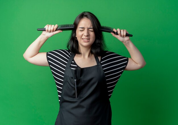 Young beautiful female hairdresser in apron with gold medal around neck holding two hair brushes hear her temples disappointed  over green