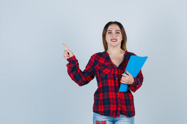 Young beautiful female holding folder, pointing at upper left corner in casual shirt and looking cheerful , front view.