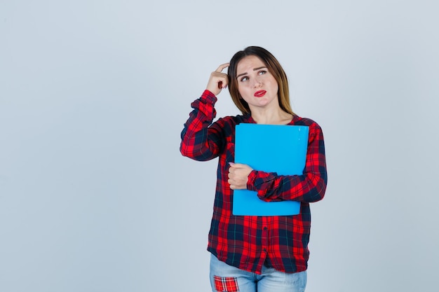 Young beautiful female holding folder, scratching her head and looking upwards in casual shirt, jeans and looking pensive. front view.