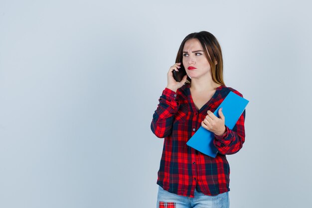 Young beautiful female holding folder while talking on phone, looking away in casual shirt, jeans and looking bewildered , front view.