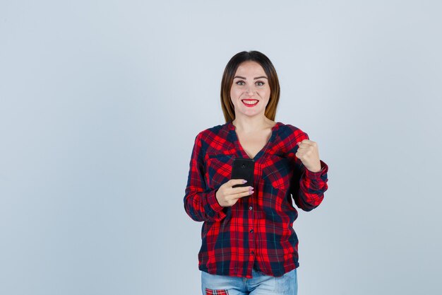 Young beautiful female holding phone, showing clenched fist in casual shirt and looking proud , front view.