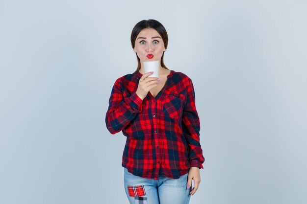 Young beautiful female holding plastic glass near mouth in casual shirt, jeans and looking curious , front view.