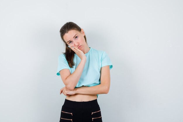 Young beautiful female leaning cheek on palm in t-shirt, pants and looking upset , front view.