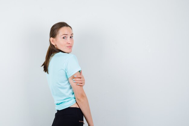 Young beautiful female looking over shoulder with hand on arm in t-shirt, pants and looking confident. front view.