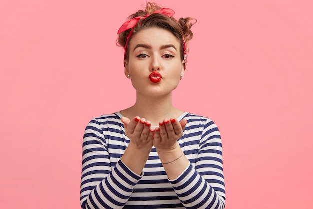 Young beautiful female model blows air kiss, expresses her love to boyfriend, wears striped sweater, isolated over pink