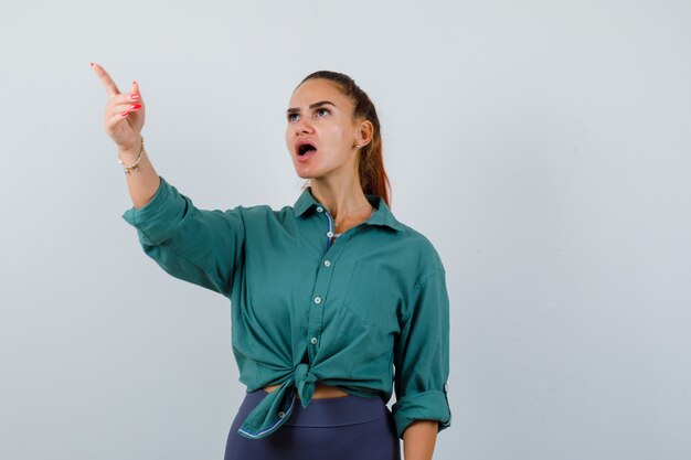 Young beautiful female pointing away in green shirt and looking shocked. front view.