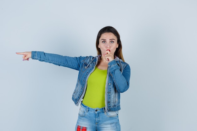 Young beautiful female pointing left while keeping hand near mouth in denim outfit and looking bewildered , front view.