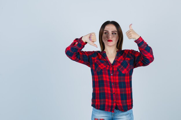 Young beautiful female pointing to opposite directions with thumbs in casual shirt and looking gloomy , front view.