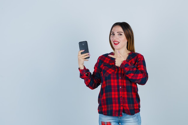 Young beautiful female pointing at phone in casual shirt and looking blissful. front view.