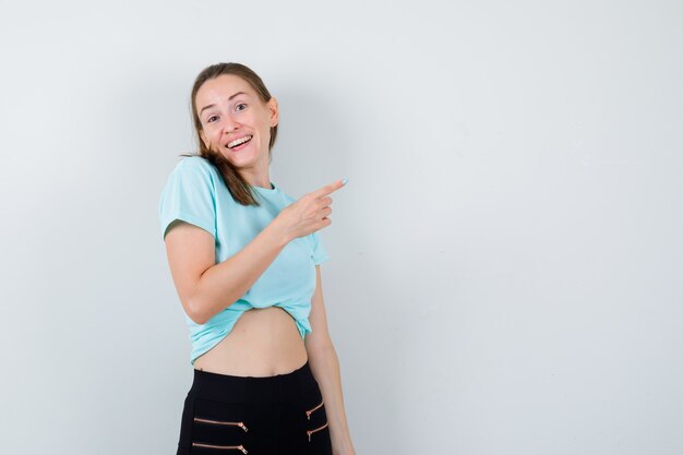 Young beautiful female pointing at upper right corner in t-shirt, pants and looking glad. front view.