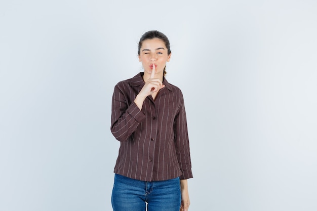 Young beautiful female showing silence gesture, winking eye in jacket and looking focused , front view.