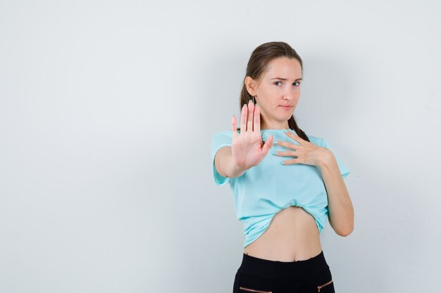 Young beautiful female showing stop gesture, with hand on chest in t-shirt and looking confident. front view.