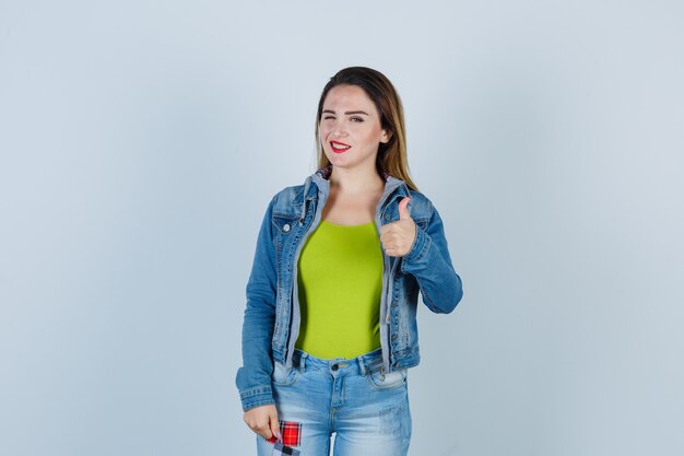 Young beautiful female showing thumb up, winking eye in denim outfit and looking cheerful. front view.