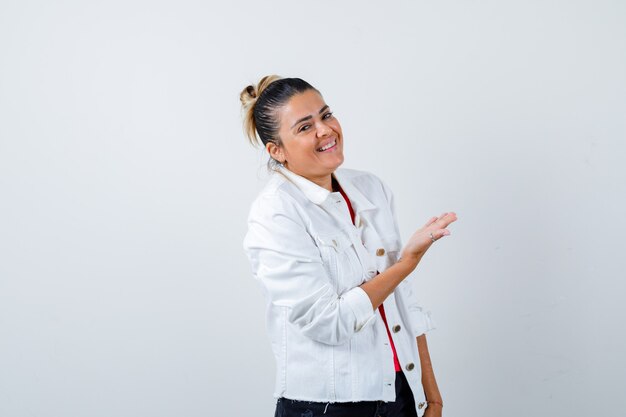 Young beautiful female showing welcome gesture in t-shirt, white jacket and looking joyous. front view.