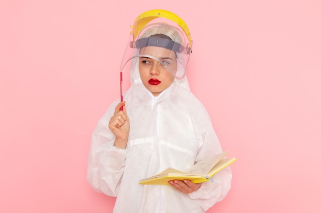 Young beautiful female in special white suit wearing protective helmet holding copybook on pink