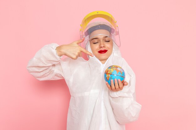 young beautiful female in special white suit and yellow helmet holding little round globe on pink