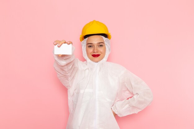 молодая красивая женщина в специальном белом костюме и желтом шлеме держит белую карточку на розовом