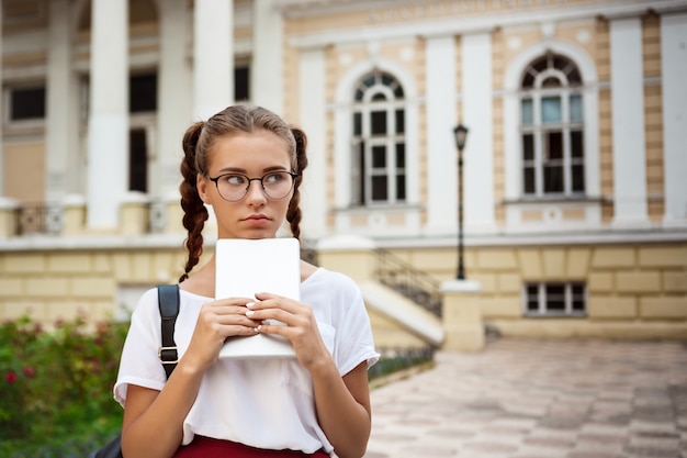 Young beautiful female student in glasses holding tablet, thinking outdoors.