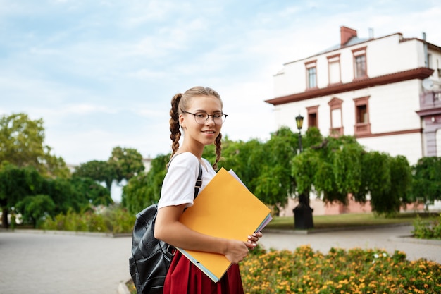 Young beautiful female student in glasses smiling, holding folders outdoors.