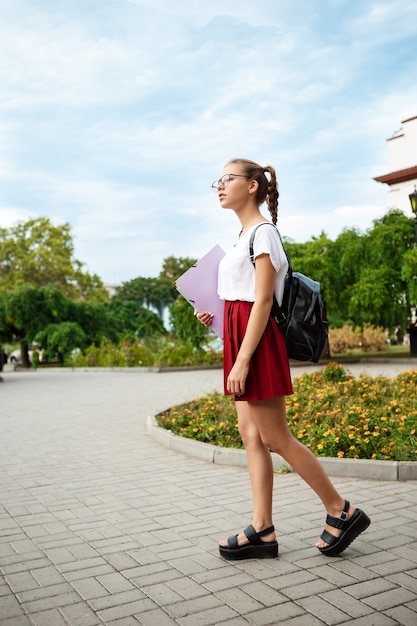 Молодая красивая студентка в очках, улыбаясь, держа папки на открытом воздухе.