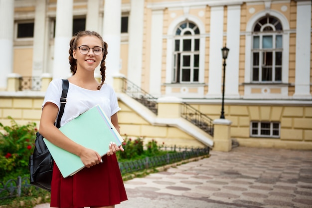 Молодая красивая студентка в очках, улыбаясь, держа папки на открытом воздухе.