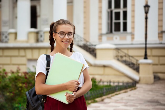 笑みを浮かべて、屋外のフォルダーを保持しているメガネの若い美しい女子学生。