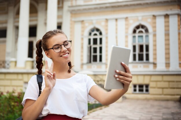 Young beautiful female student in glasses smiling, making selfie outdoors.